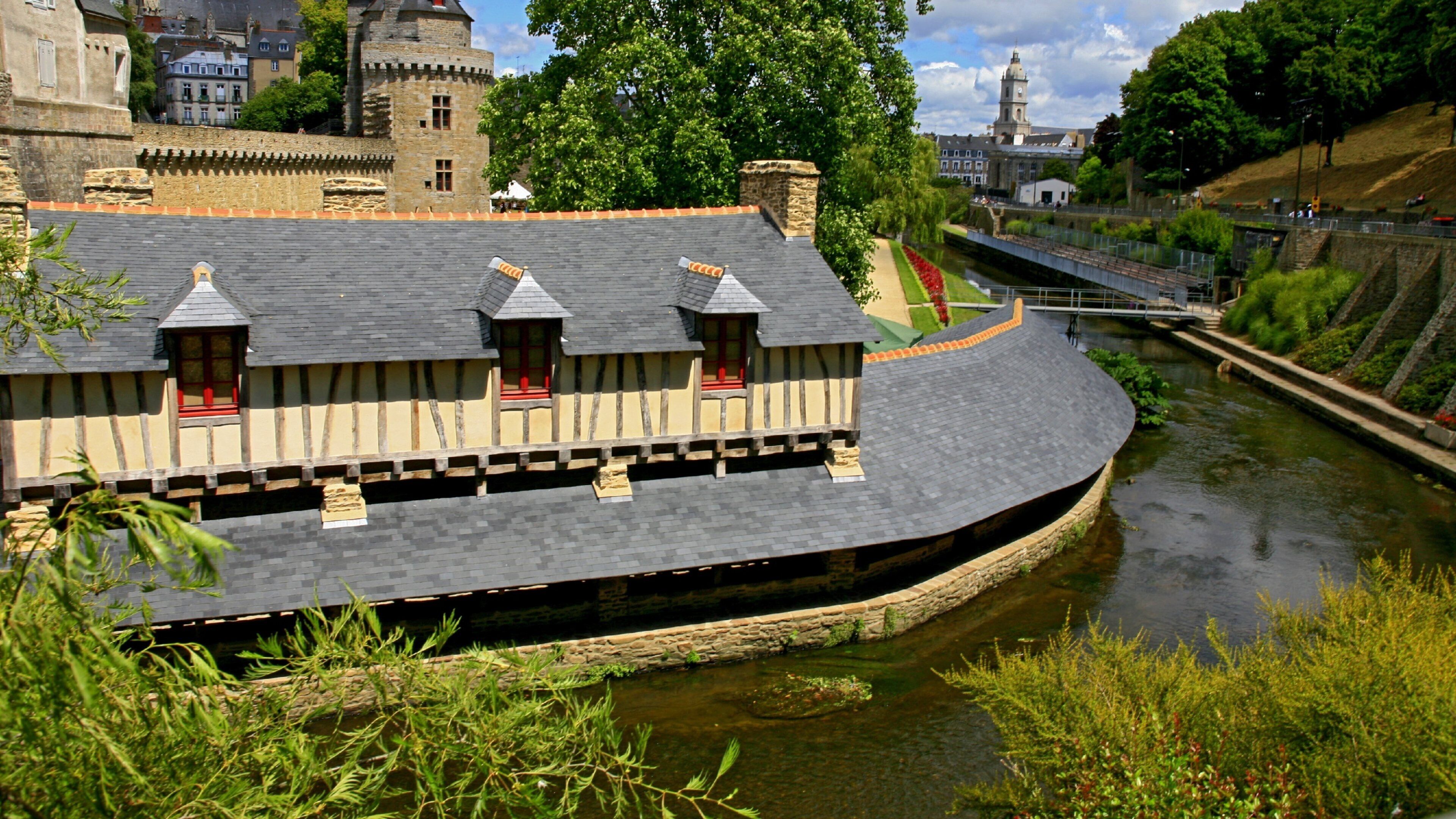 A riverside building with a curved roof, a stone wall, and a bridge in the background.