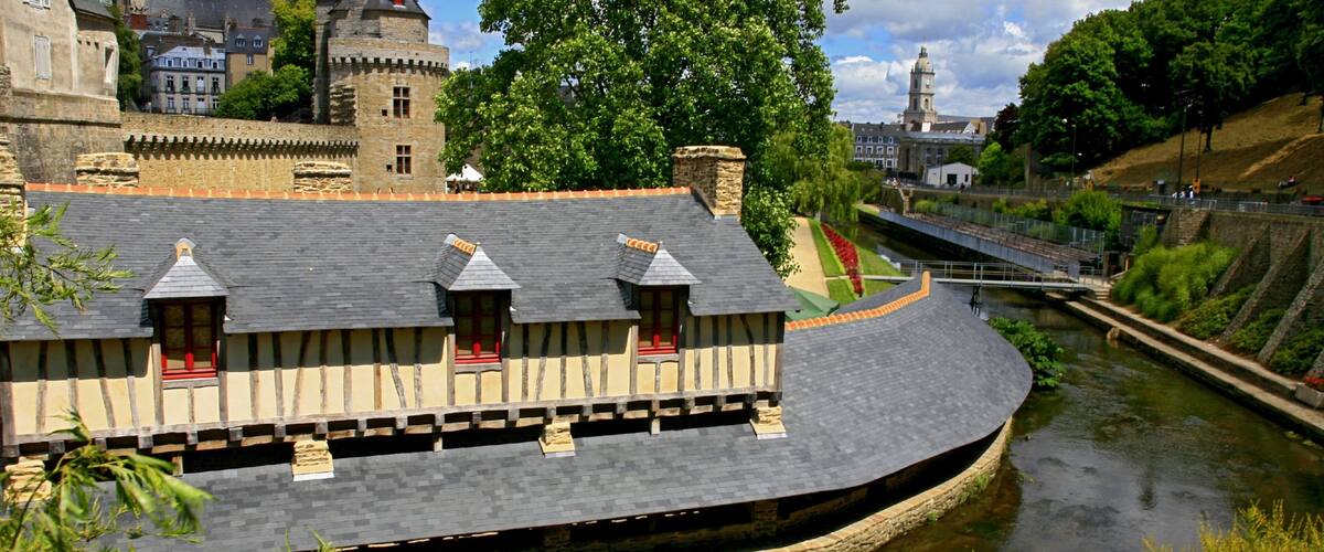 A riverside building with a curved roof, a stone wall, and a bridge in the background.