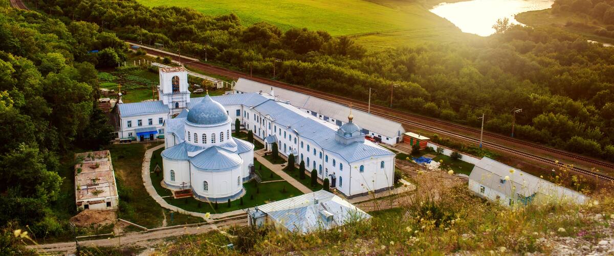 A large white church with blue domes surrounded by lush greenery and a river.