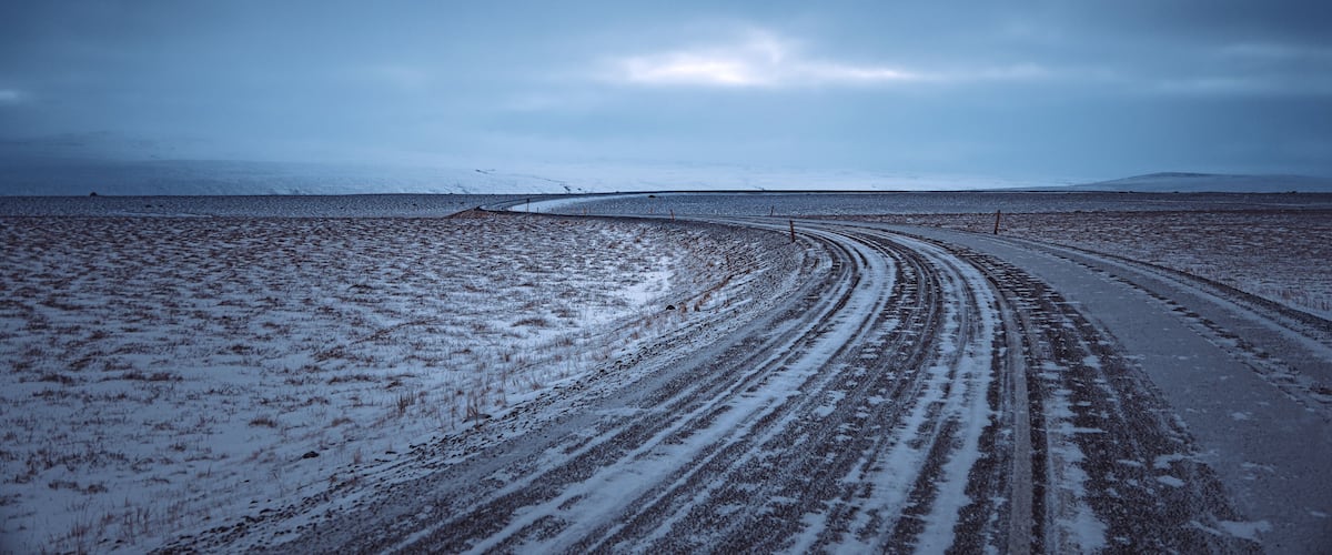 A snow-covered landscape with a curved dirt road and distant mountains.