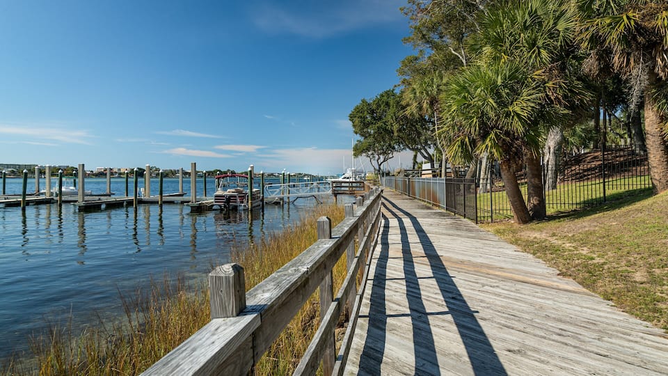 A wooden boardwalk along a calm water body with boats, trees, and a distant cityscape.