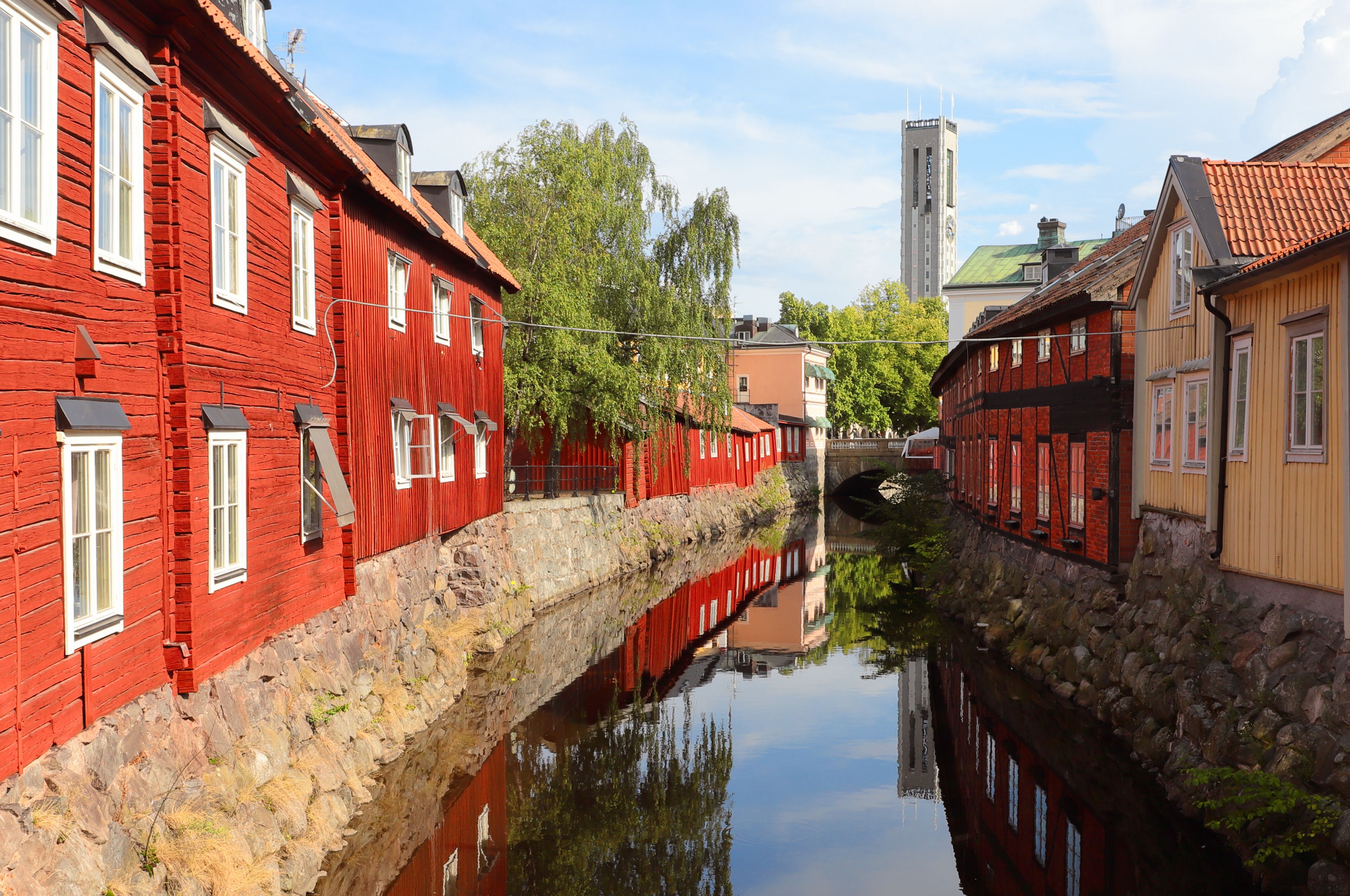 A narrow canal with red buildings on one side and a yellow building on the other, reflecting in the water.