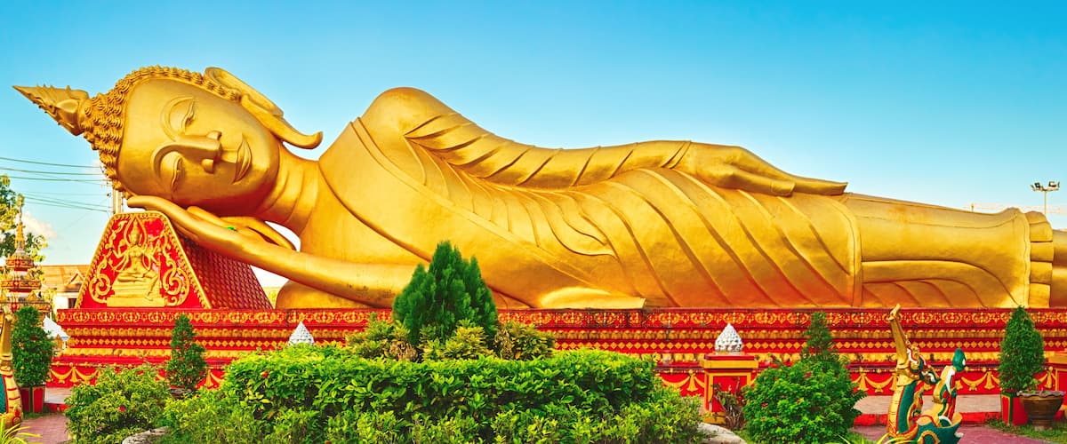 A large golden reclining Buddha statue surrounded by lush greenery and traditional architecture.