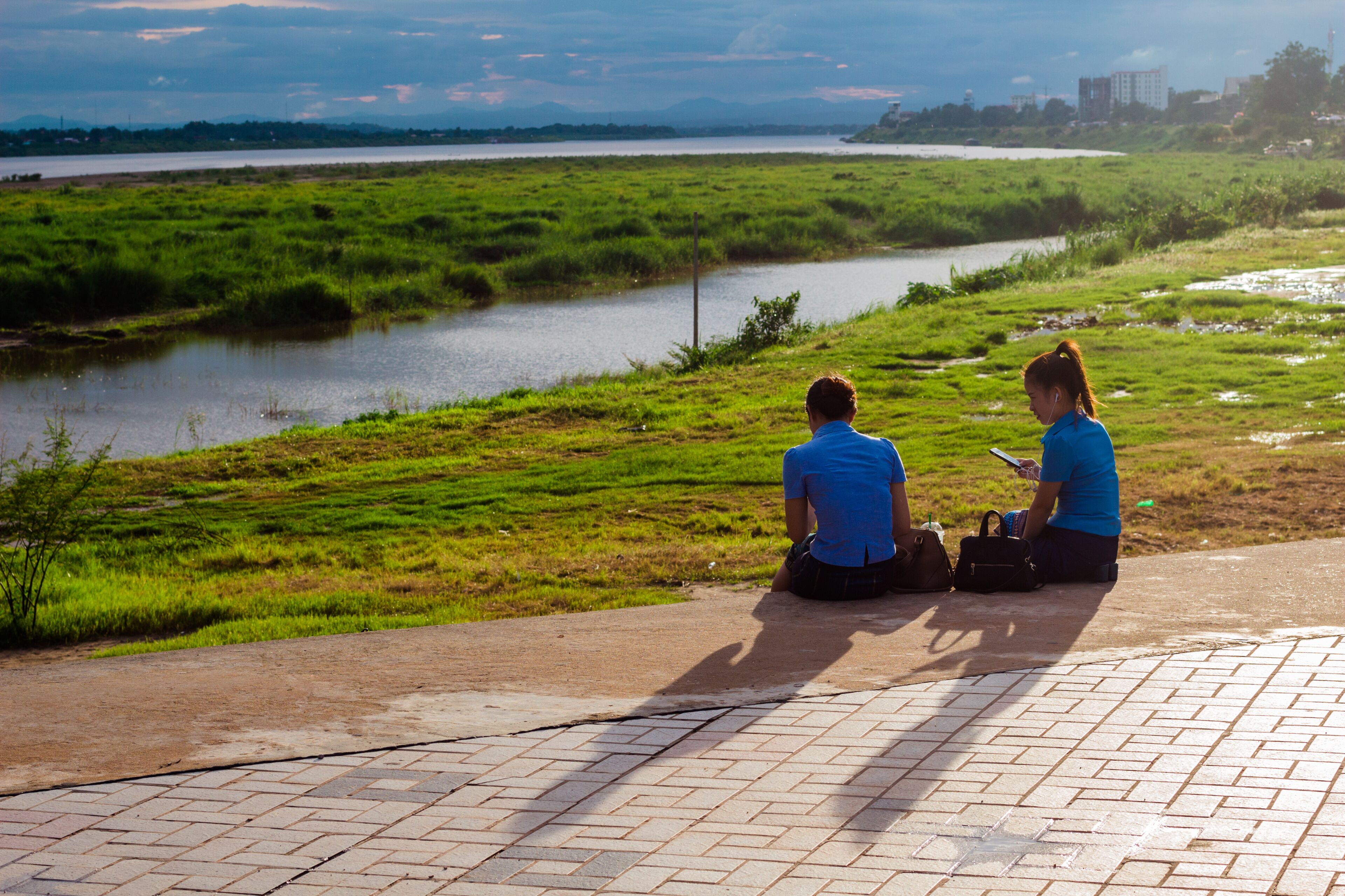 Dos personas sentadas en una zona pavimentada con vista a un río, vegetación y edificios a lo lejos.