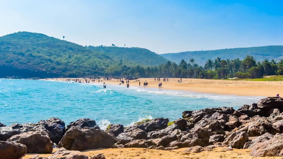 A beach with rocky foreground, clear blue water, and hills in the background.
