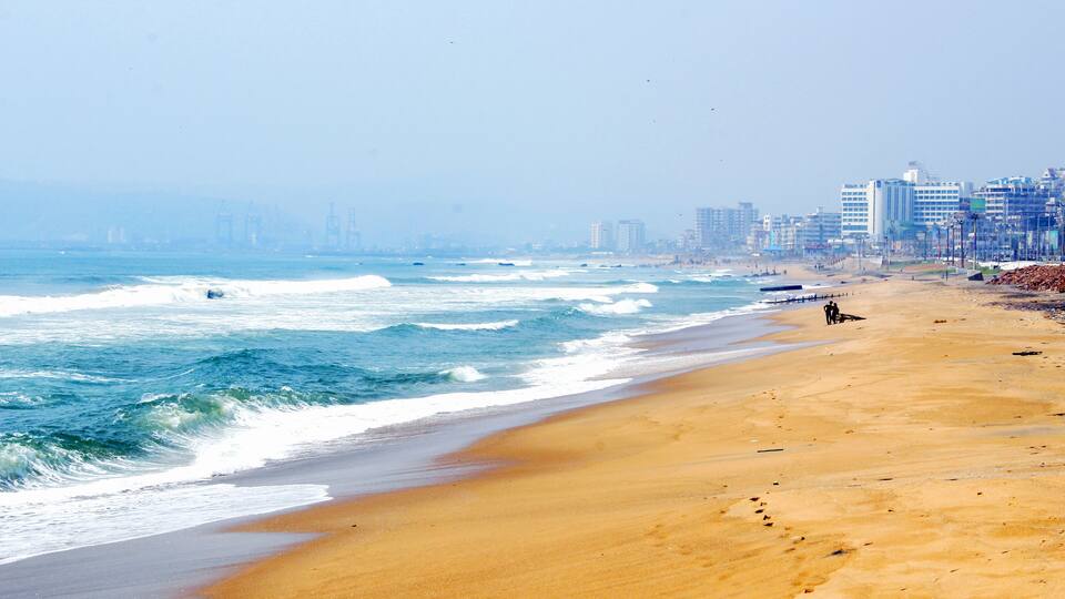 A beach with waves, a sandy shore, and buildings in the background.