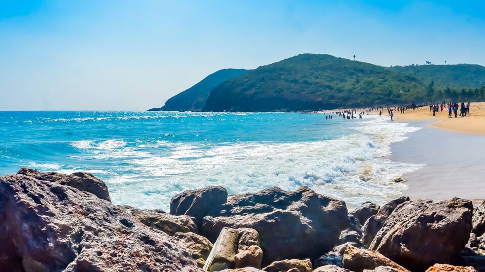 A beach with rocky foreground, clear blue water, and a hill in the background.
