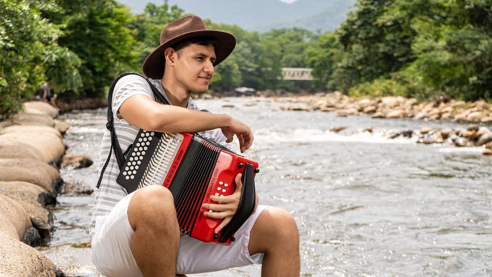 A person playing an accordion by a river with rocks and greenery in the background.