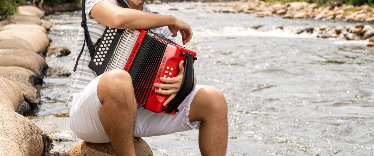 A person playing an accordion by a river with rocks and greenery in the background.