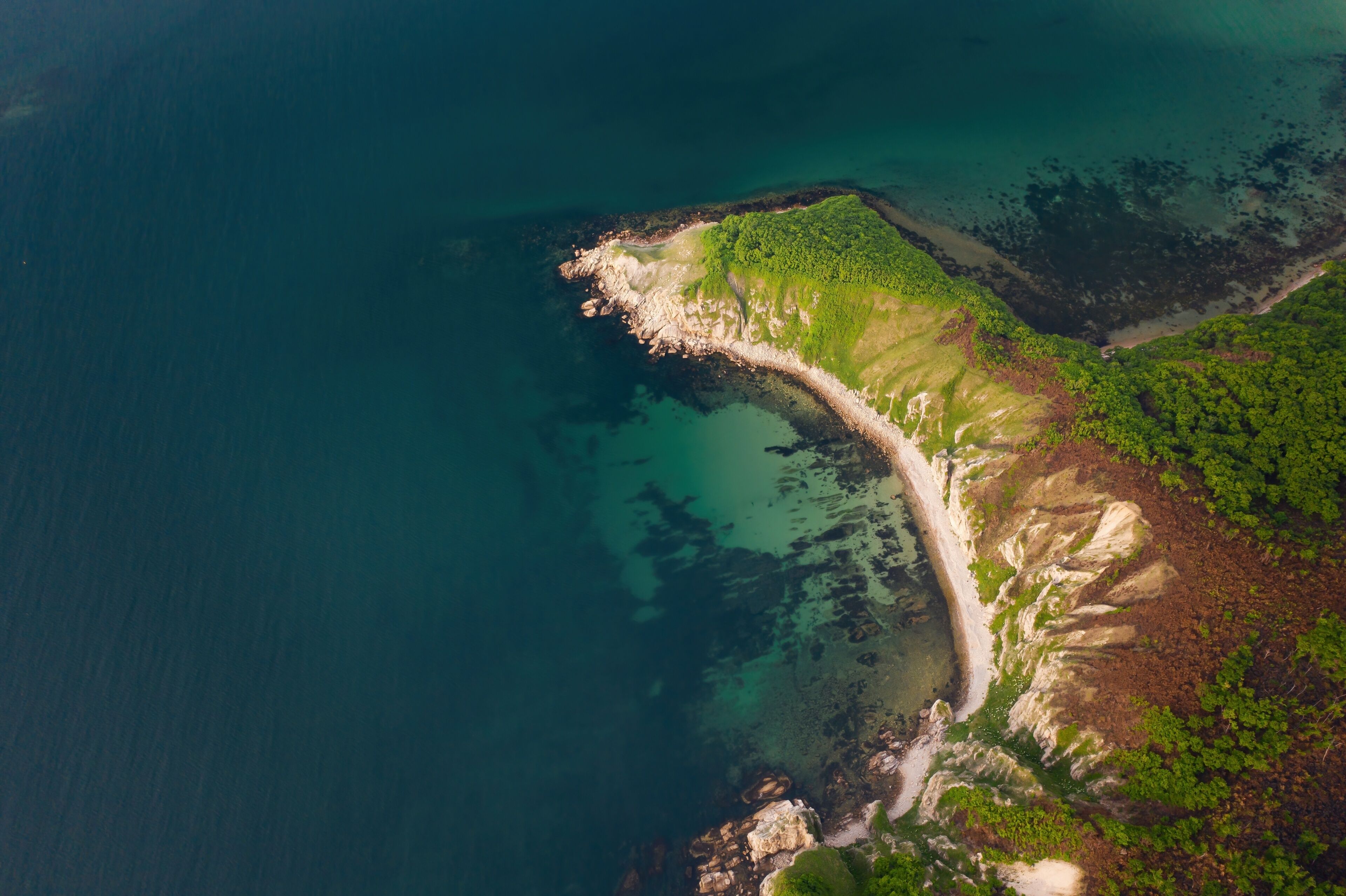 An aerial view of a coastal landscape with a sandy beach, green vegetation, and clear blue waters.