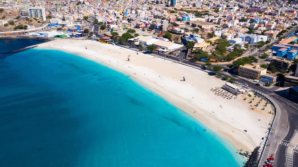A coastal city with a wide sandy beach, a clear blue sea, and densely packed buildings.