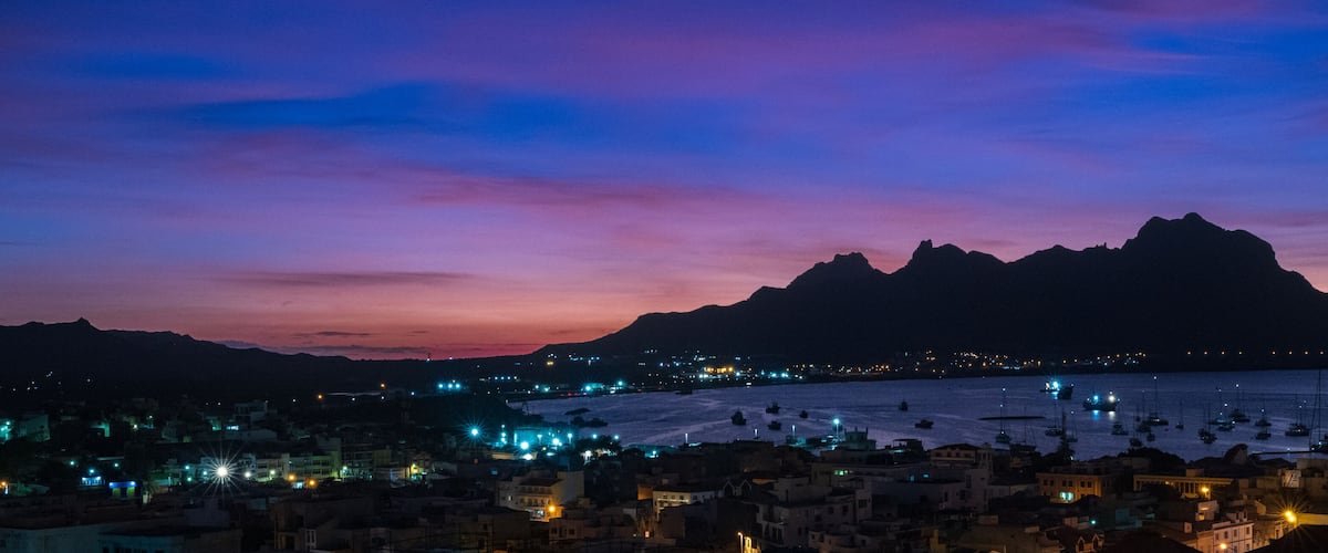 A coastal town at dusk with a mountain in the background and boats in the water.