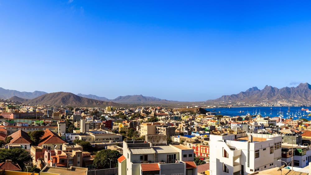 Un paysage urbain côtier avec un port de plaisance, des montagnes en arrière-plan et un ciel bleu dégagé.