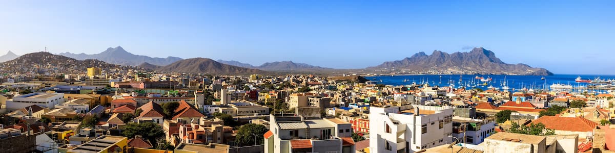 A coastal cityscape with a marina, mountains in the background, and a clear blue sky.