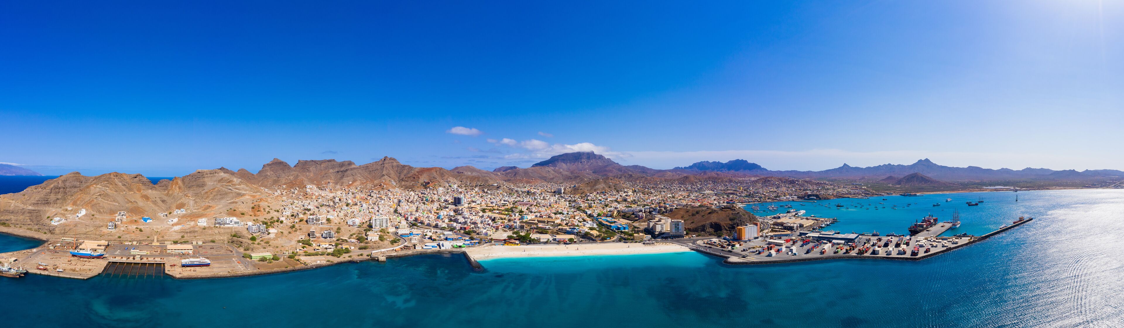 Una ciudad costera con un puerto, montañas al fondo y aguas azul turquesa.