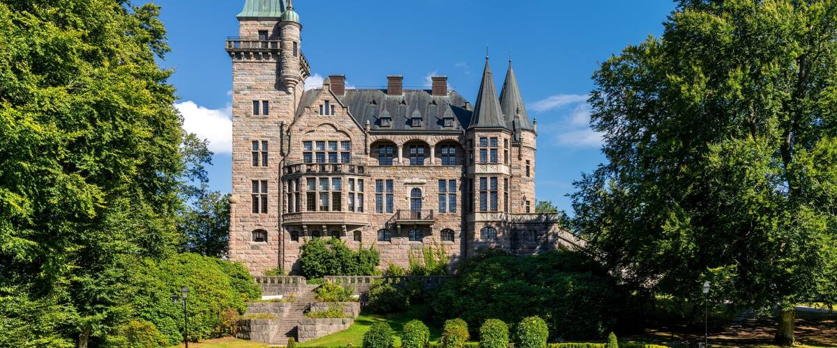 A large, historic castle with multiple towers, surrounded by well-maintained gardens and pathways.