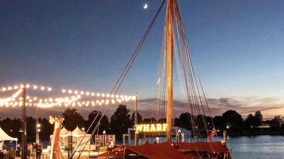 A wooden boat with a red canopy and the word "WHARF" on its side, docked at a waterfront with string lights in the background.