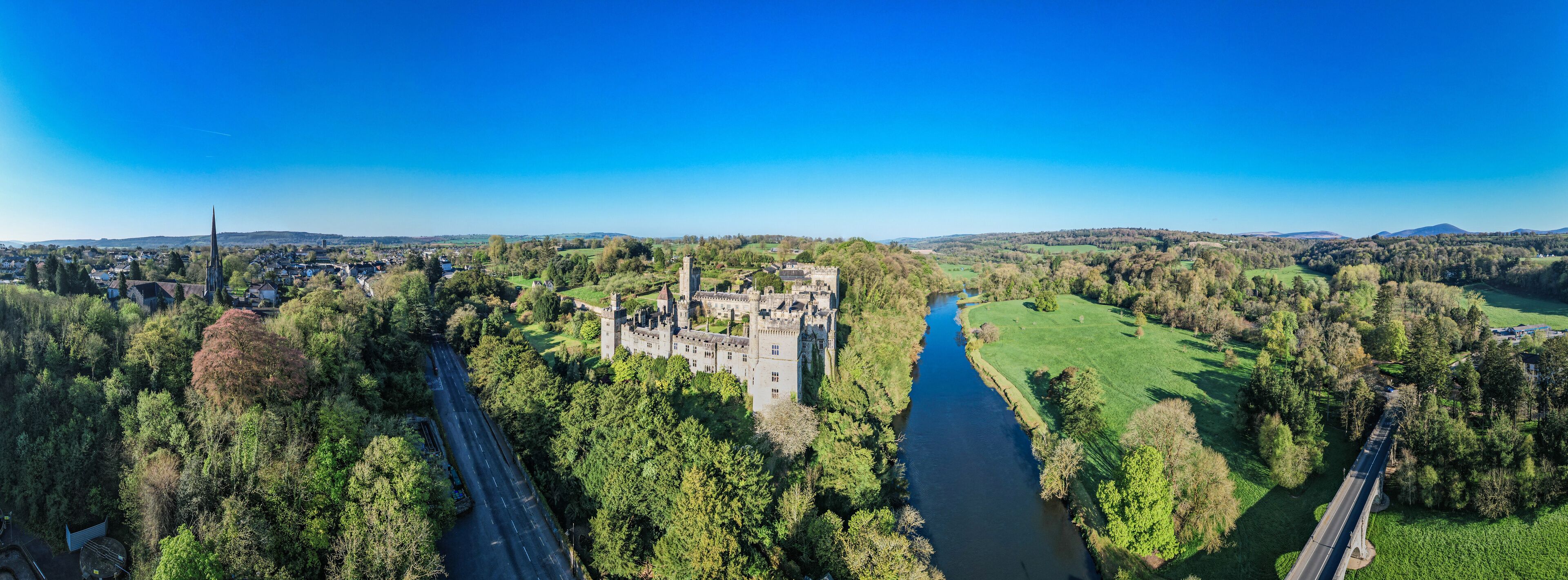 A castle by a river with surrounding greenery and a clear blue sky.