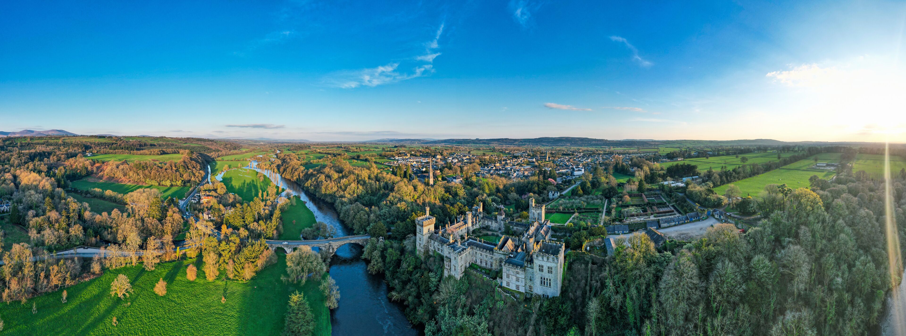 A castle surrounded by a forest and fields, with a river running through the landscape.