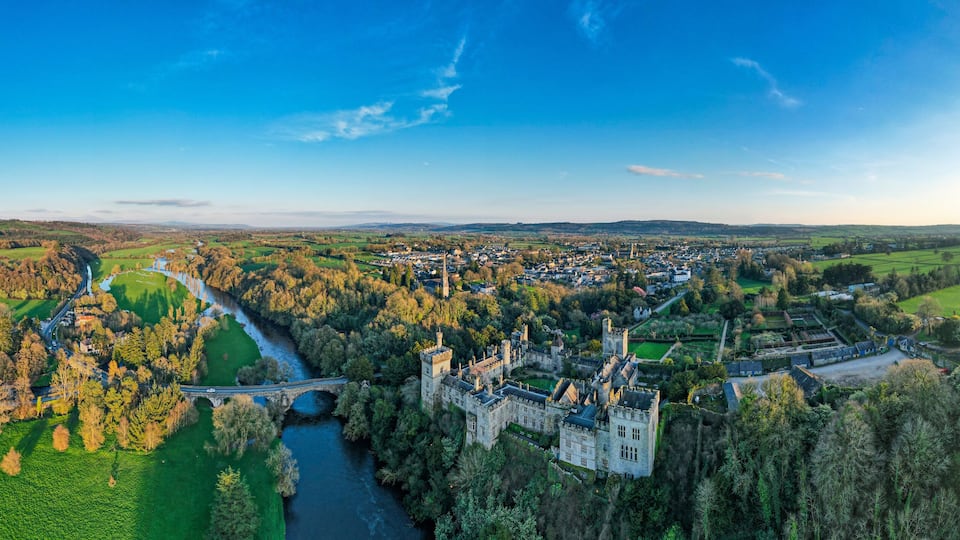 A castle surrounded by a forest and fields, with a river running through the landscape.
