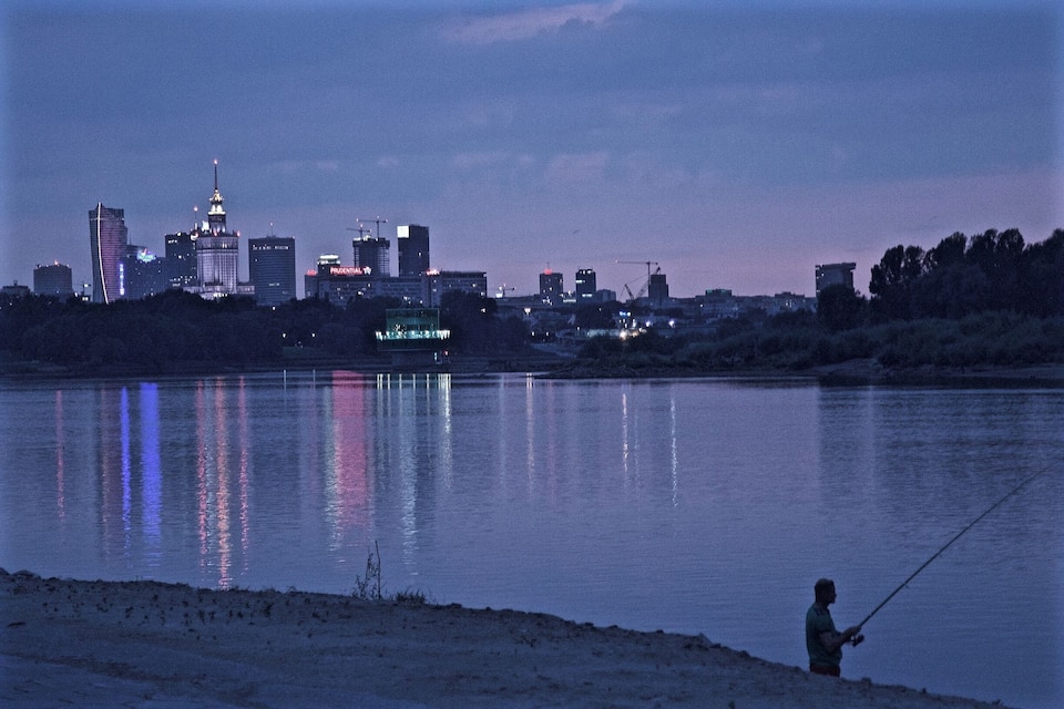 A city skyline at dusk with a person fishing by the water.