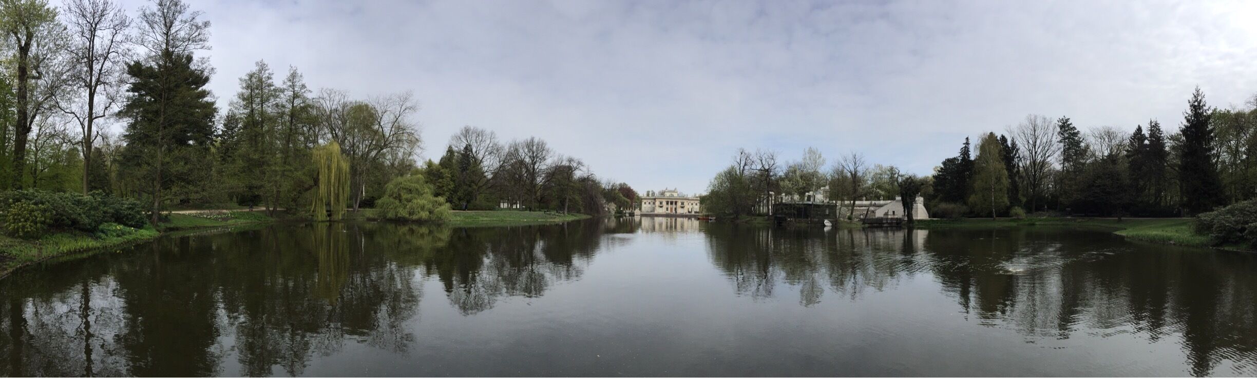 A calm river with reflections of trees and buildings, surrounded by greenery and a cloudy sky.