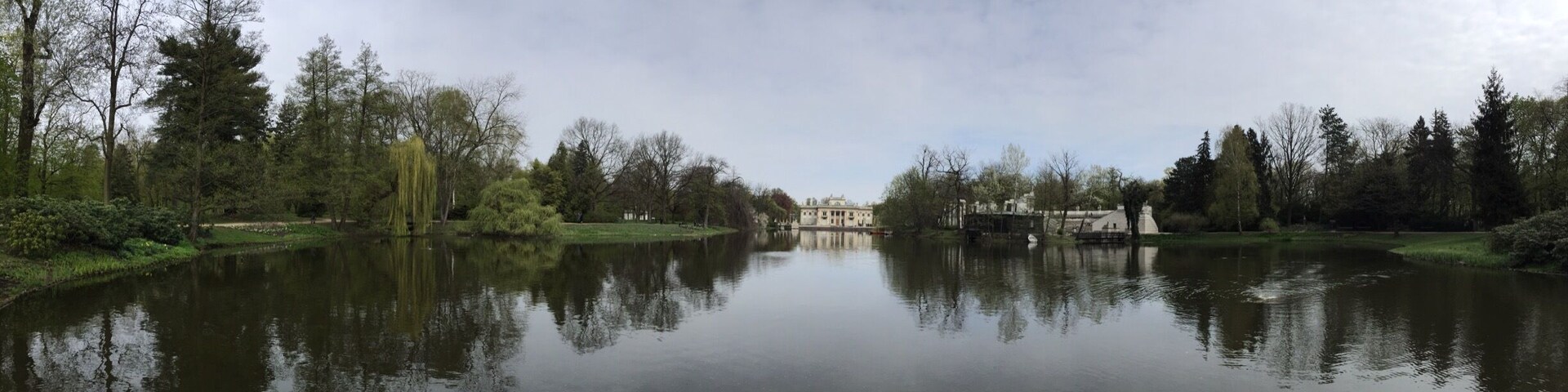 A calm river with reflections of trees and buildings, surrounded by greenery and a cloudy sky.