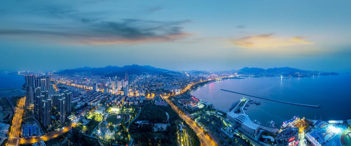 A coastal cityscape at dusk with high-rise buildings, a marina, and a large body of water.