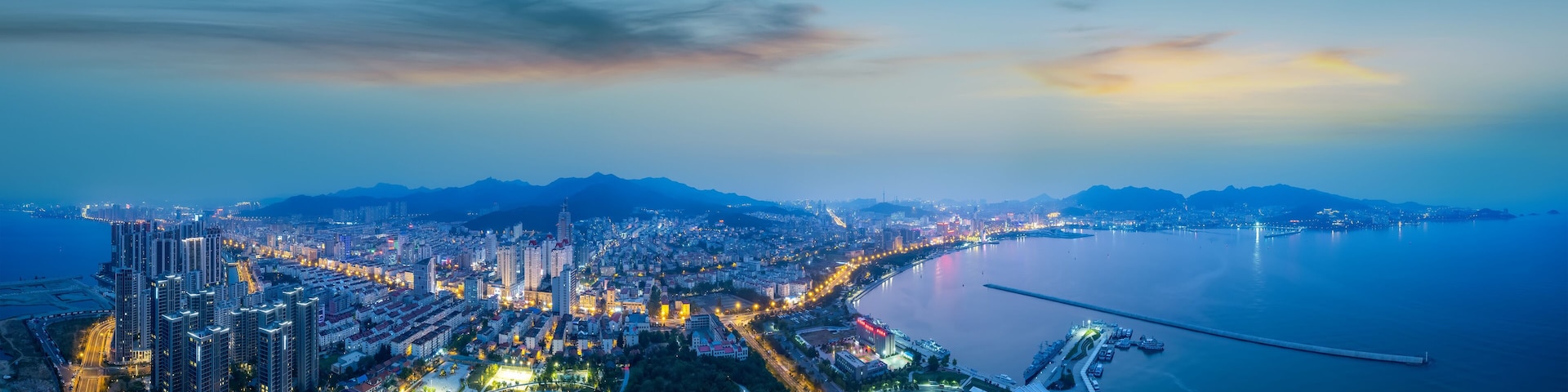 A coastal cityscape at dusk with high-rise buildings, a marina, and a large body of water.