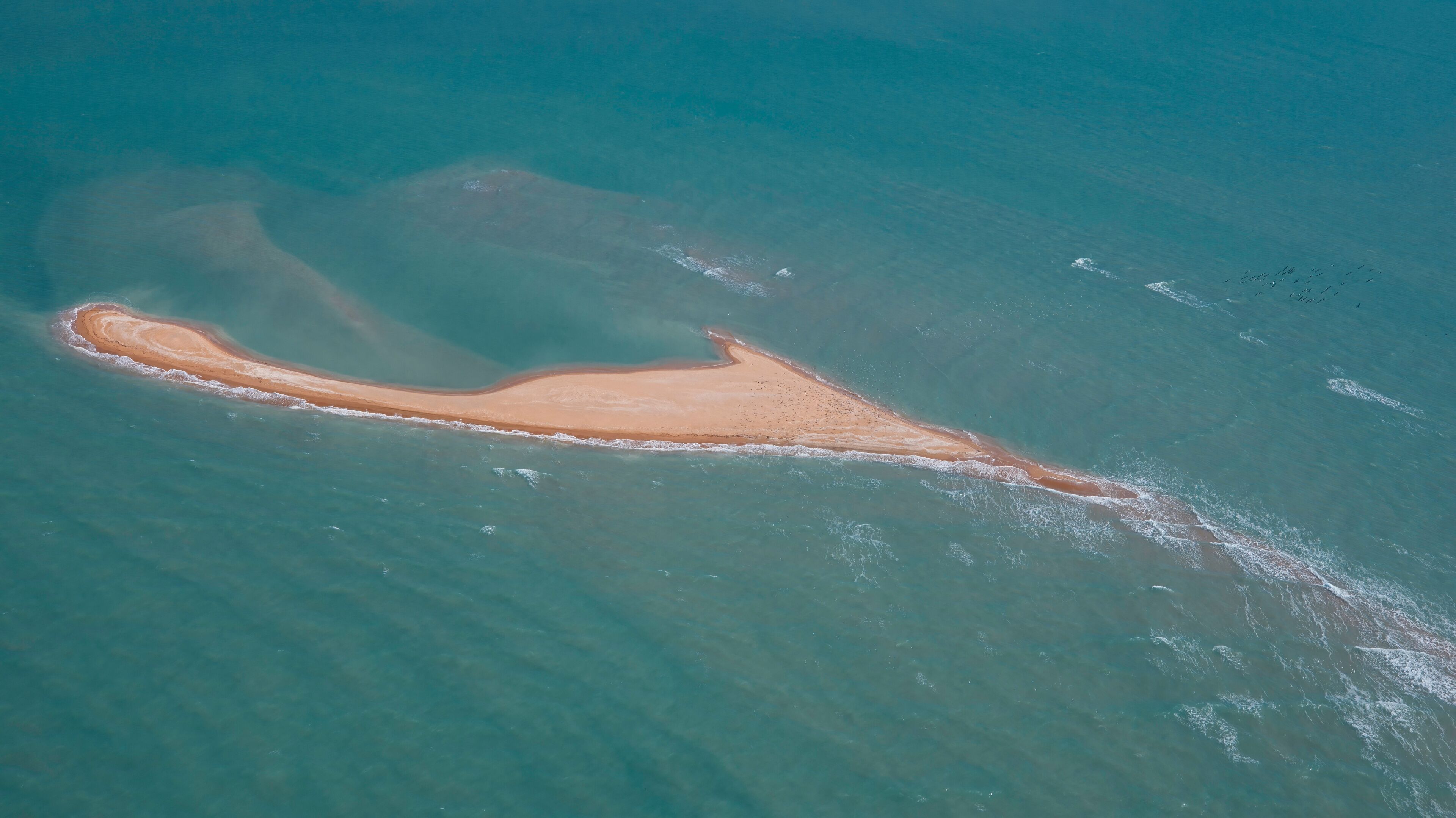 An aerial view of a narrow, elongated island with a sandy beach and clear blue waters.