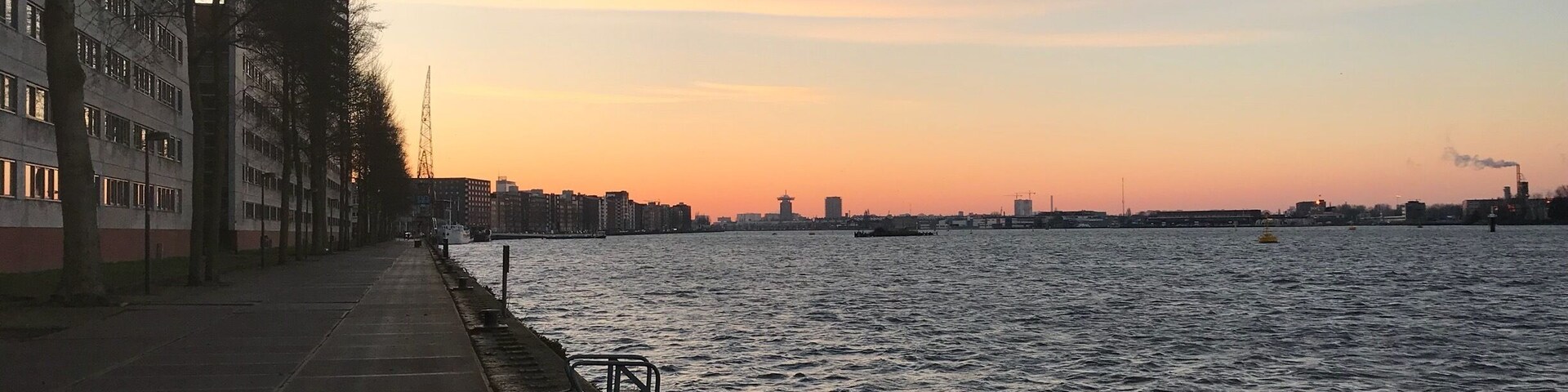 Ein Uferweg mit Gebäuden an einer Seite und einem Sonnenuntergang über dem Wasser.