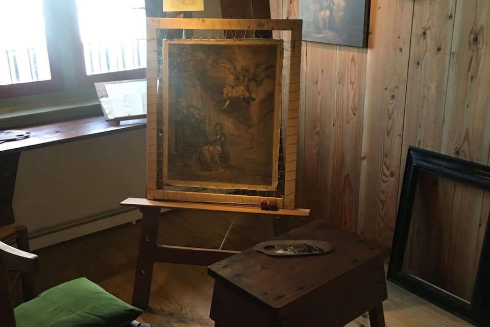 A rustic room with a wooden floor, a green cushioned chair, an easel with a painting, and a framed picture on the wall.