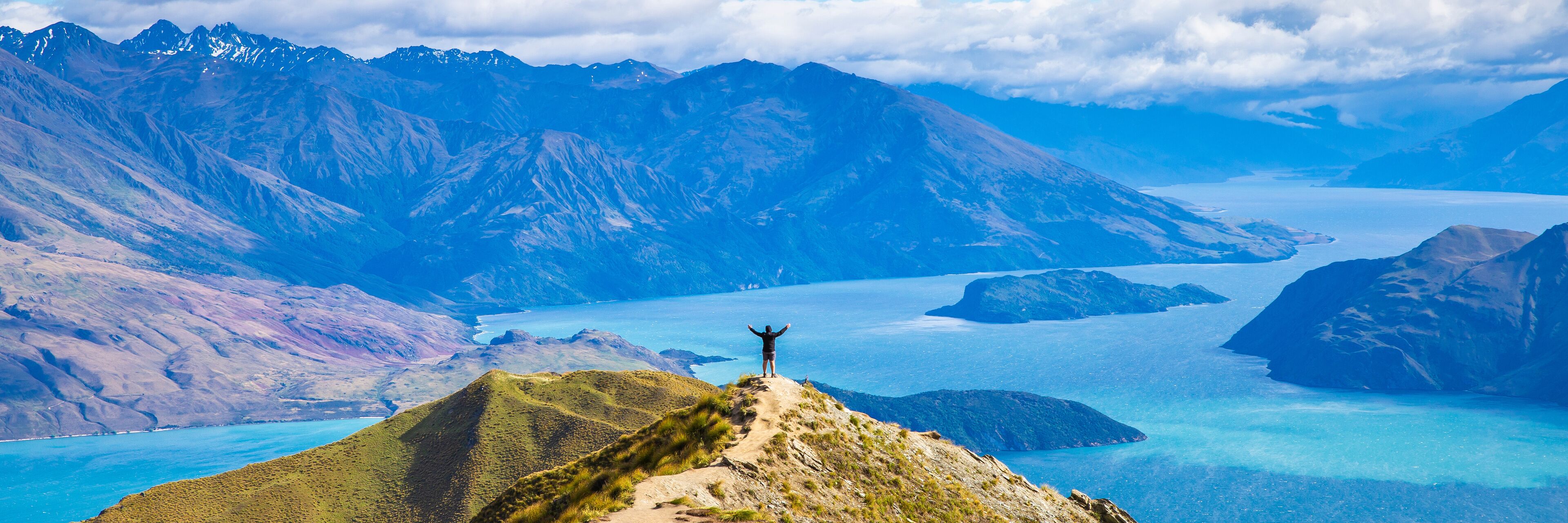 A person standing on a mountain peak with arms raised, overlooking a large body of water surrounded by rugged mountains.