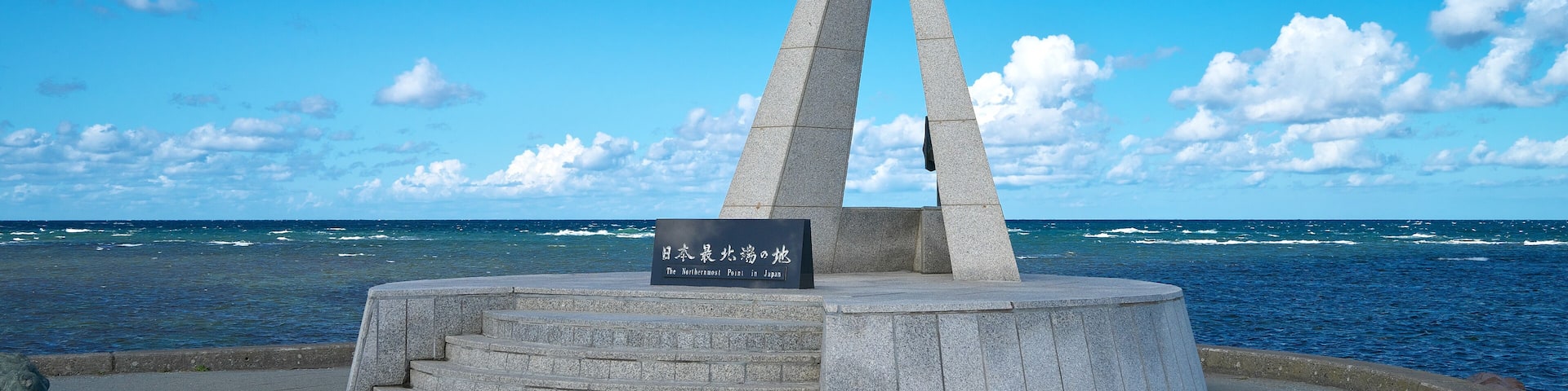 A monument with a sign in Japanese, located by the sea with a clear sky.