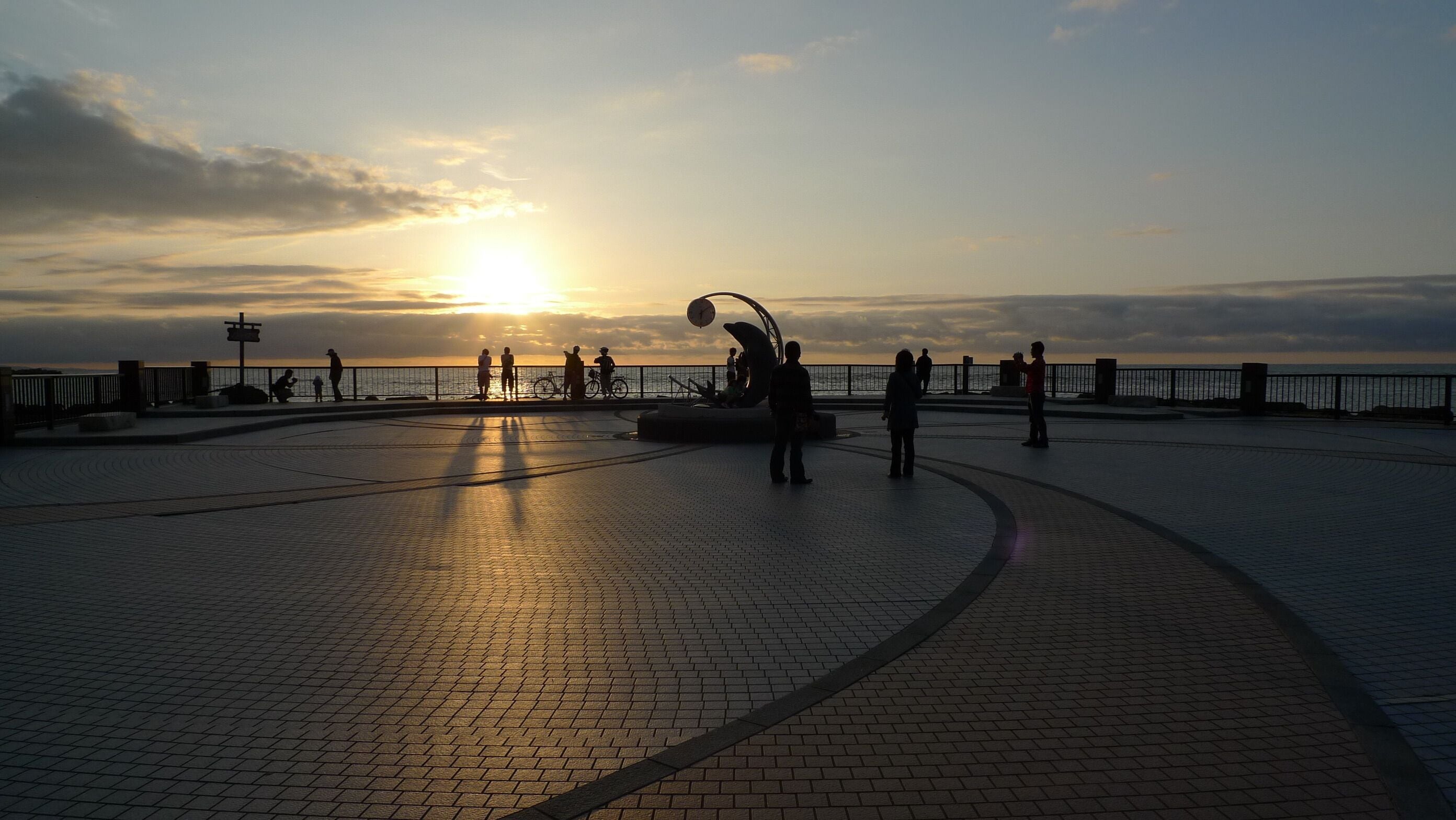 A sunset over a waterfront promenade with people walking and a sculpture in the center.