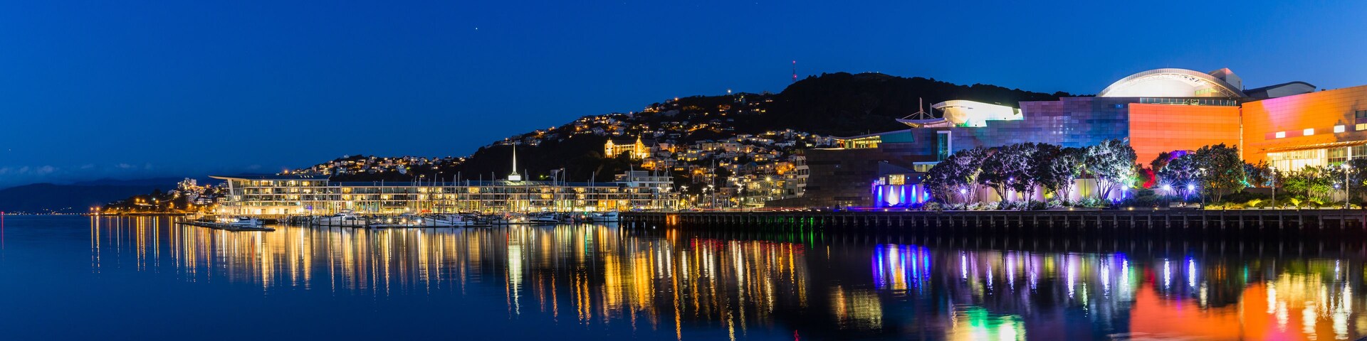 Paisagem urbana à beira-mar à noite, com edifícios iluminados, um cais e uma colina com luzes.