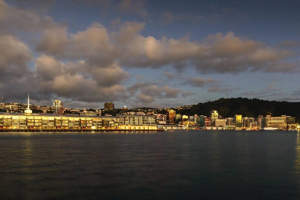 Une vue sur une ville côtière au crépuscule, avec des bâtiments illuminés et une tour imposante.