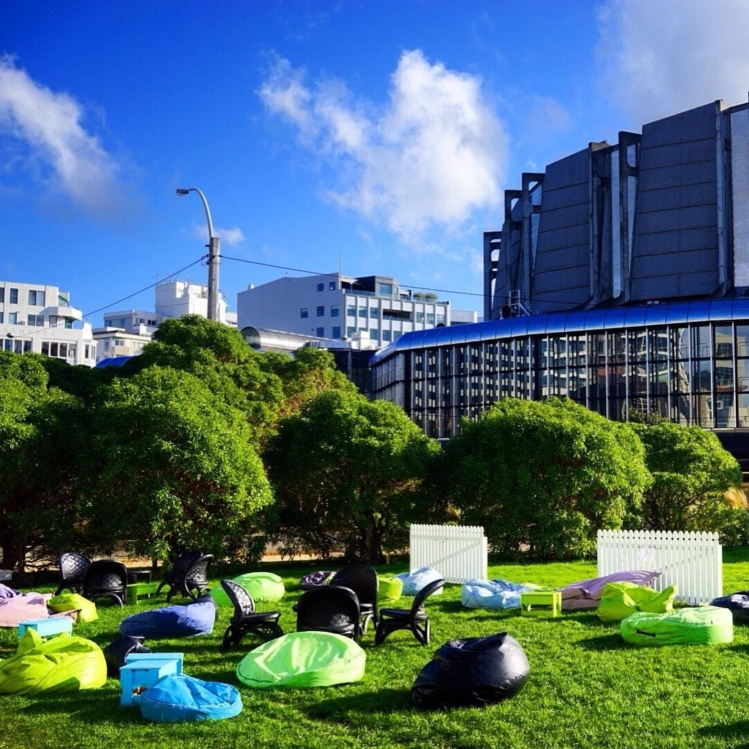Um parque com grama verde, almofadas coloridas e edifícios modernos ao fundo.