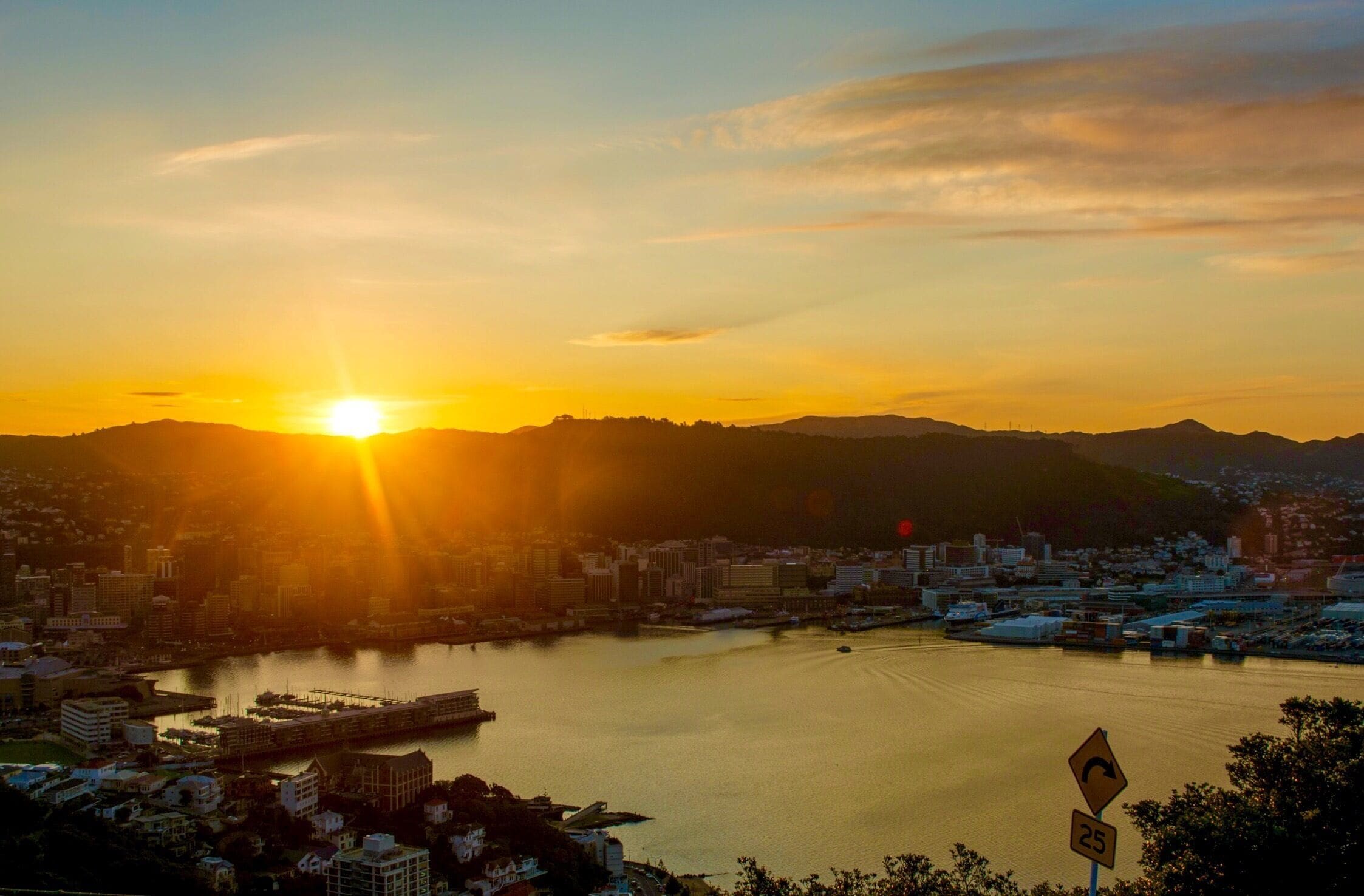 Atardecer sobre una ciudad con un río, montañas al fondo y una señal de límite de velocidad.
