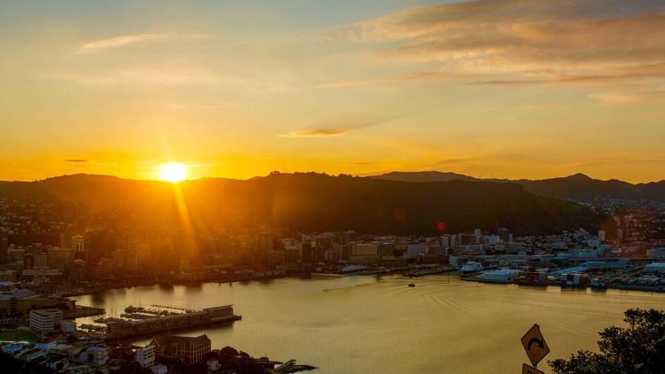 Atardecer sobre una ciudad con un río, montañas al fondo y una señal de límite de velocidad.