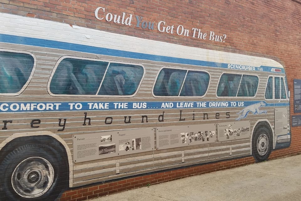 Un mural d'un autocar Greyhound sobre un mur de maons, amb text i fotografies històriques.