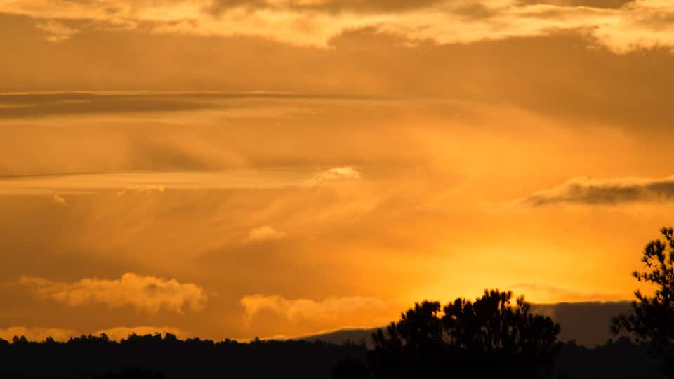 A sunset over a mountainous landscape with silhouettes of trees and a cloudy sky.