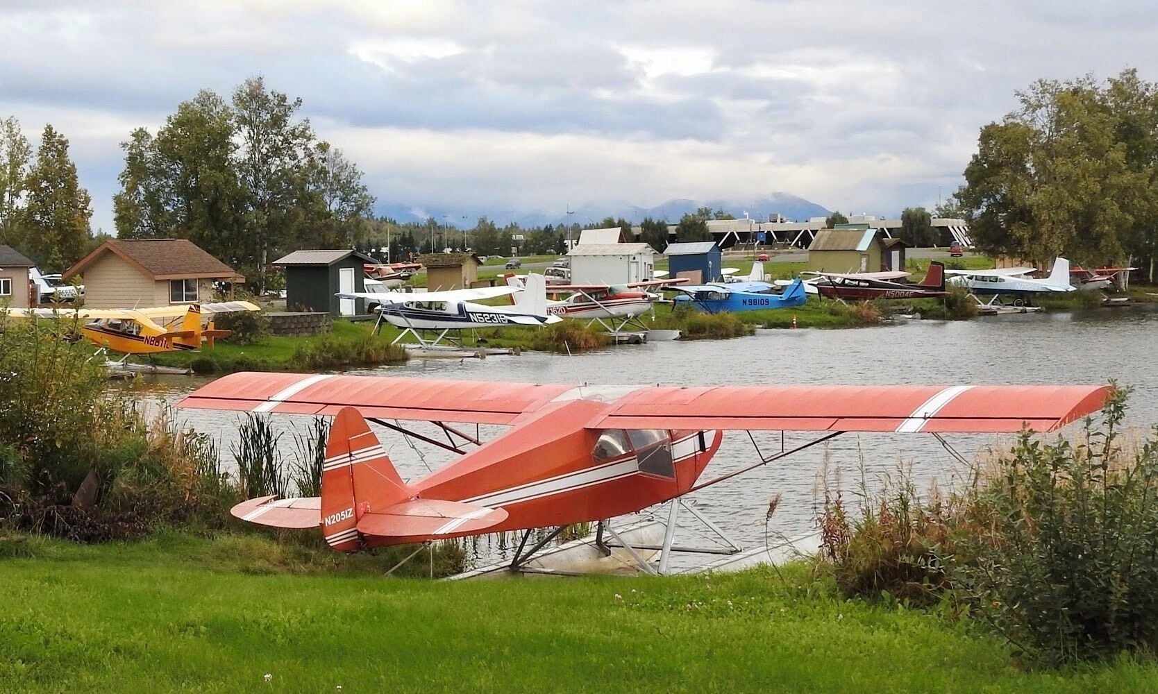 A red biplane parked near a lake with other aircraft and houses in the background.