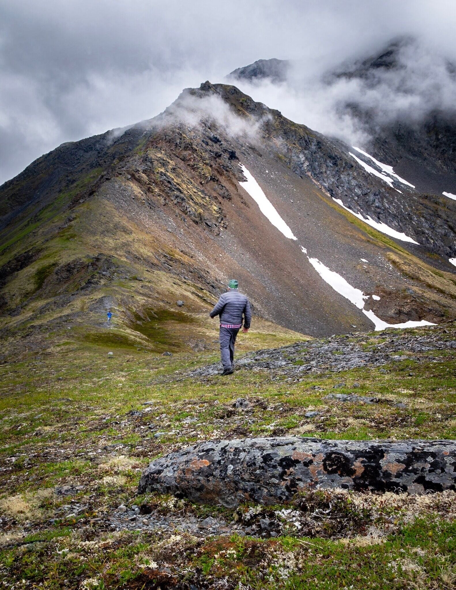 A hiker on a rocky mountain path with patches of snow and clouds.