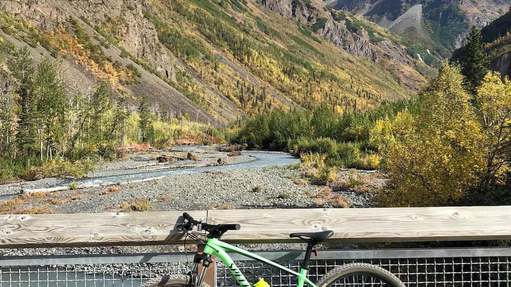 A green mountain bike parked on a wooden railing overlooking a river and mountains.