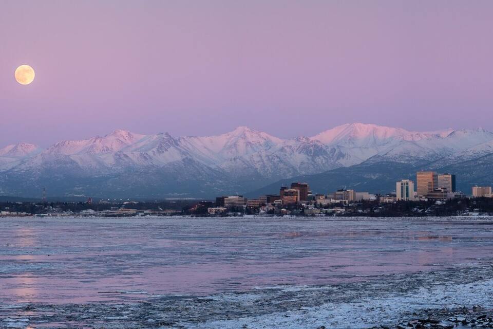 A cityscape with snow-covered mountains in the background and a frozen river in the foreground.
