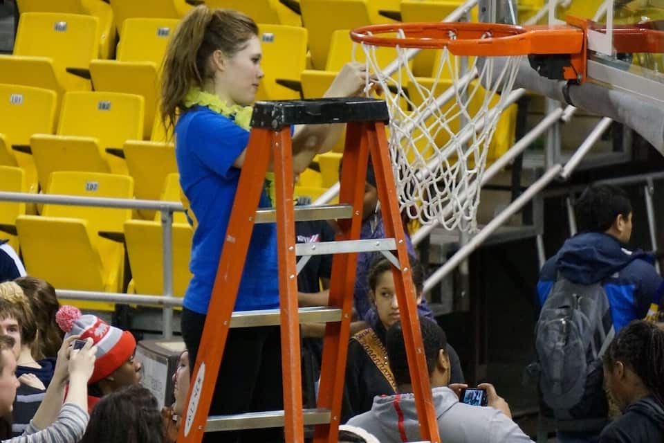 Um jogador de basquete em uma escada perto da cesta, torcedores ao redor e arquibancadas amarelas ao fundo.