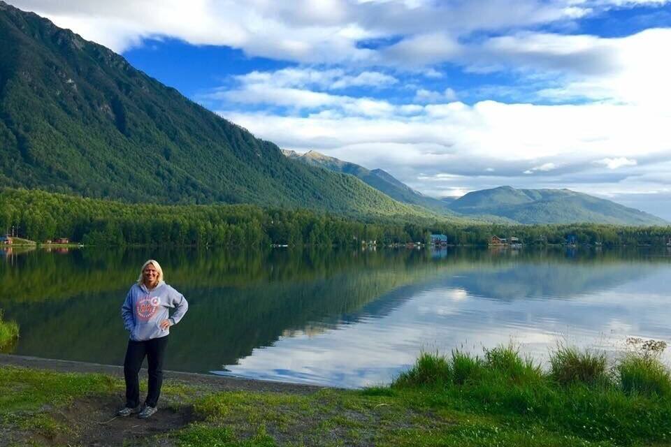 A person standing by a calm lake with mountains in the background.
