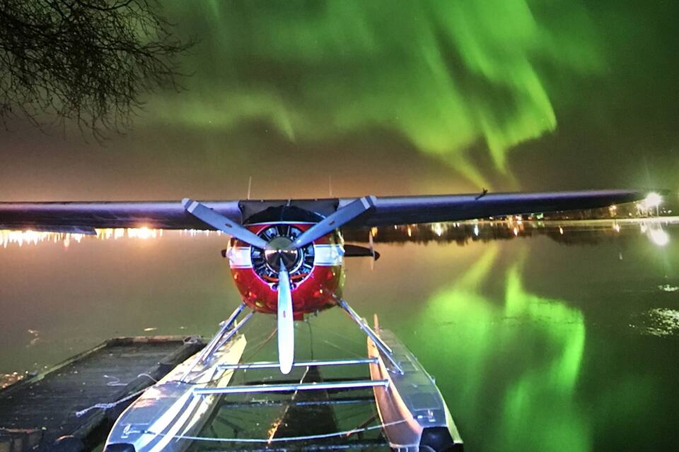 A seaplane on a calm water body with reflections of lights and a green aurora in the background.