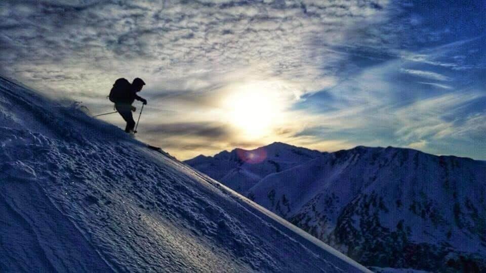 A hiker on a snowy mountain slope with a sun setting in the background.